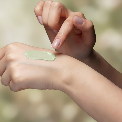 Hand applying a green cream to another hand against a blurred natural background